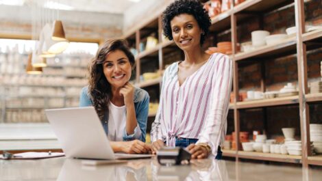 Two women smile at the camera with a laptop and a card reader on the table and shelves in the background.