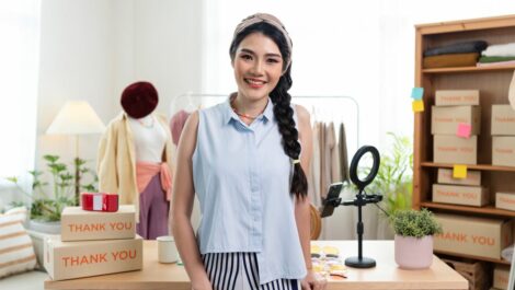 A young small business owner smiles in front of a desk with shipping boxes behind.