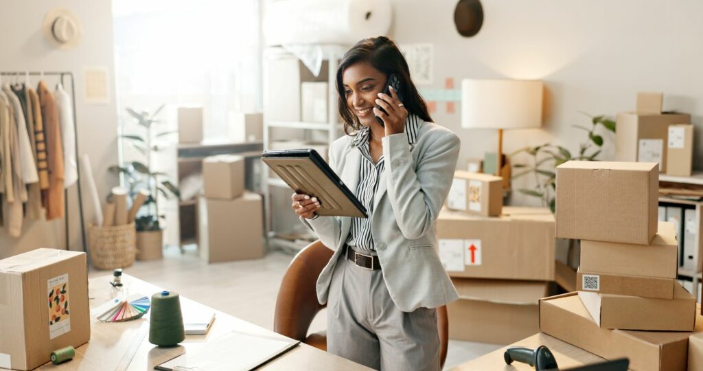 A young small business owner talking on the phone and looking at a tablet with shipping boxes behind her.