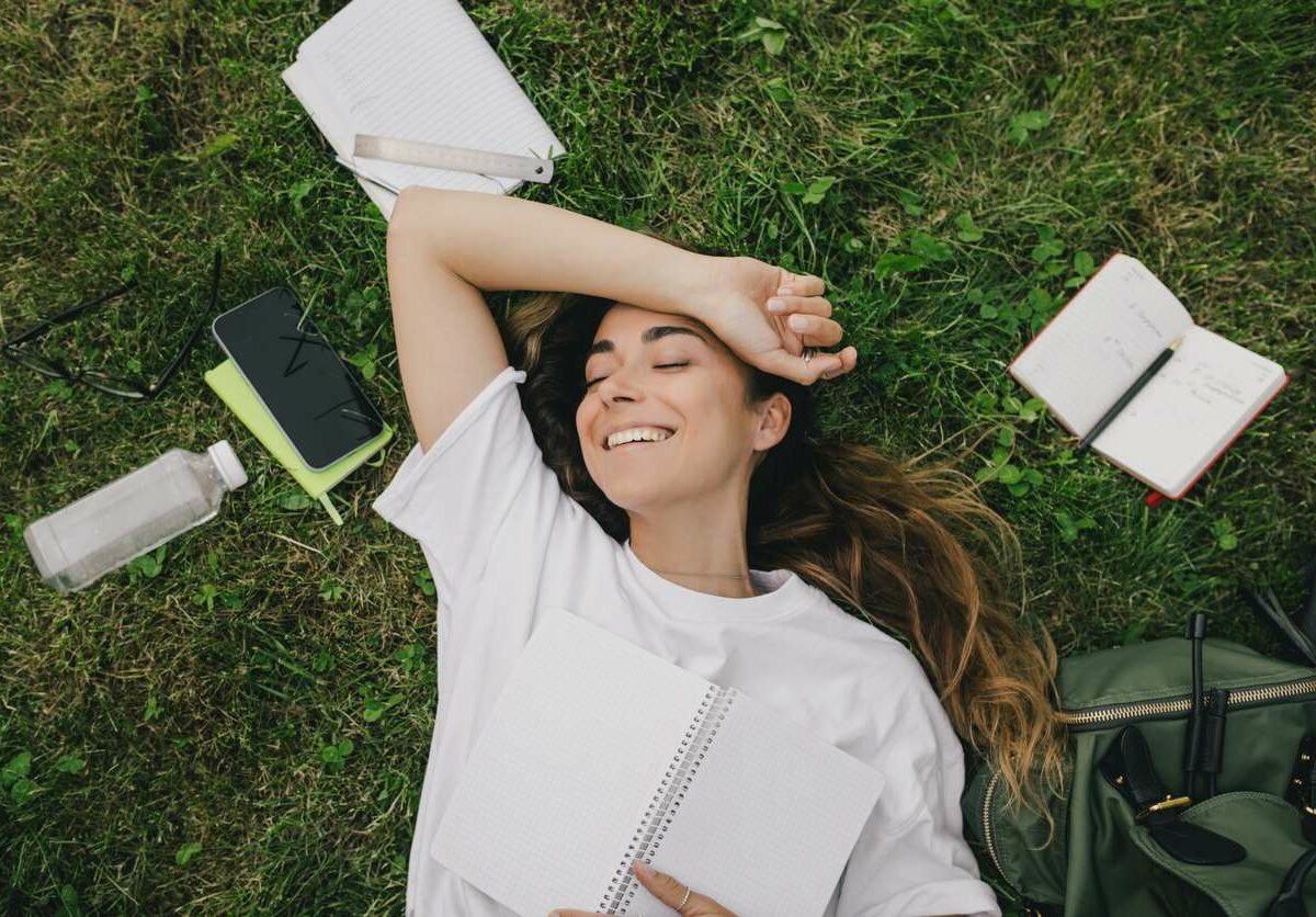 A young woman takes a break from studying using her books, laptop, and notebook while sitting on the grass.