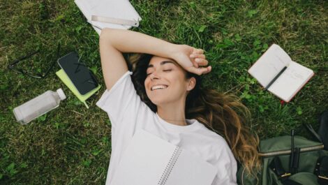 A young woman takes a break from studying using her books, laptop, and notebook while sitting on the grass.